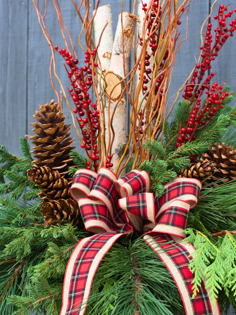 Decorative arrangement with greenery, red berries, pinecones, and a plaid ribbon against a wooden background.