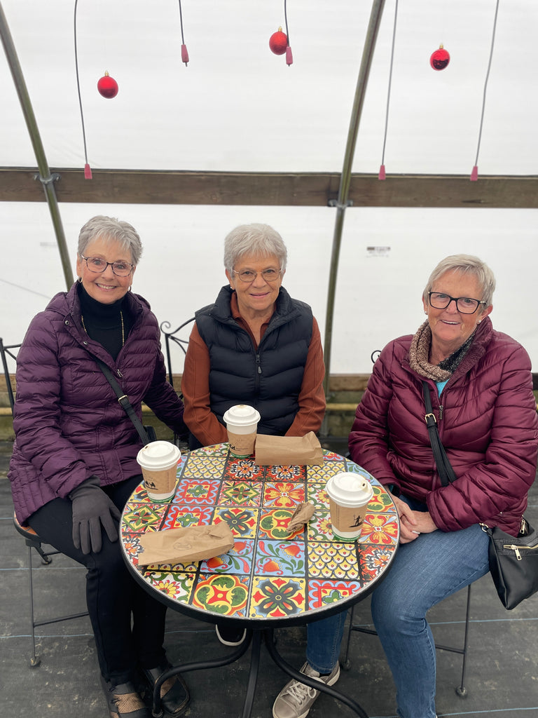 Three women sitting at a colourful table with coffee cups, wearing winter coats at Open House.